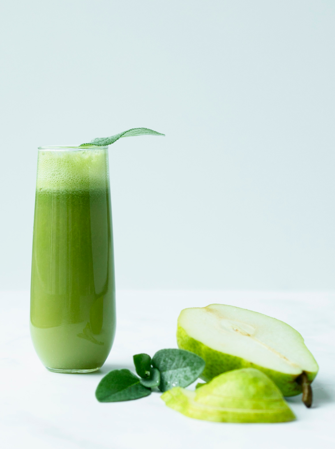 Glass filled with rich green Pear Bellini mocktail and half a pear, sliced apple and mint leaves on the side.